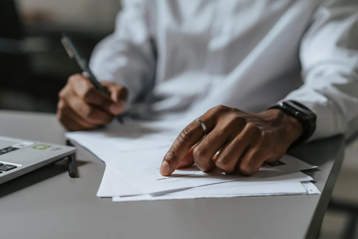 Person writing on paperwork beside an open laptop