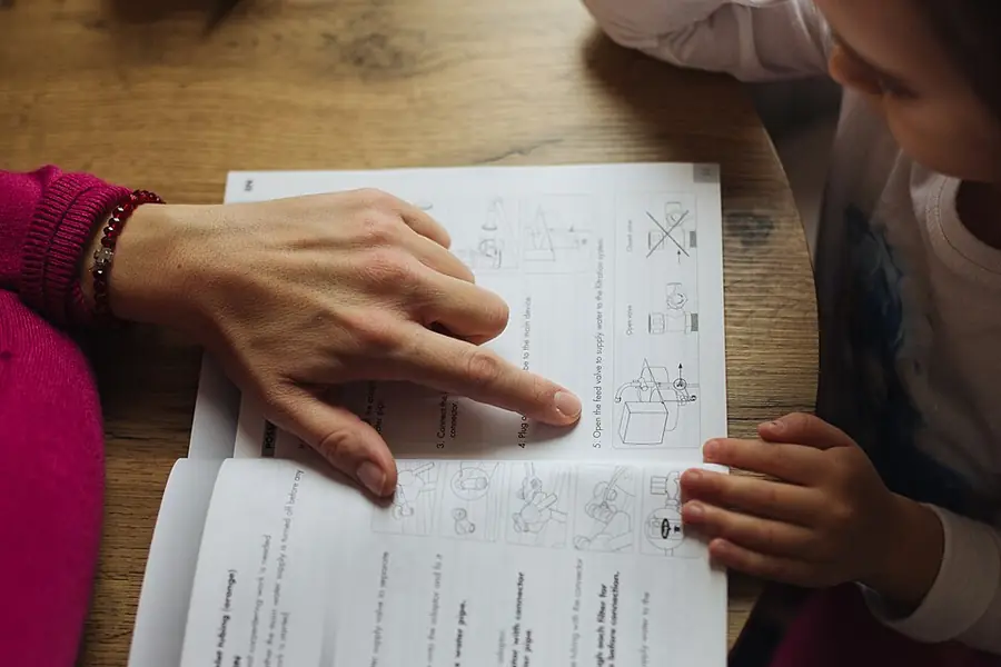 Parent points to a workbook while a child follows along at a wooden table